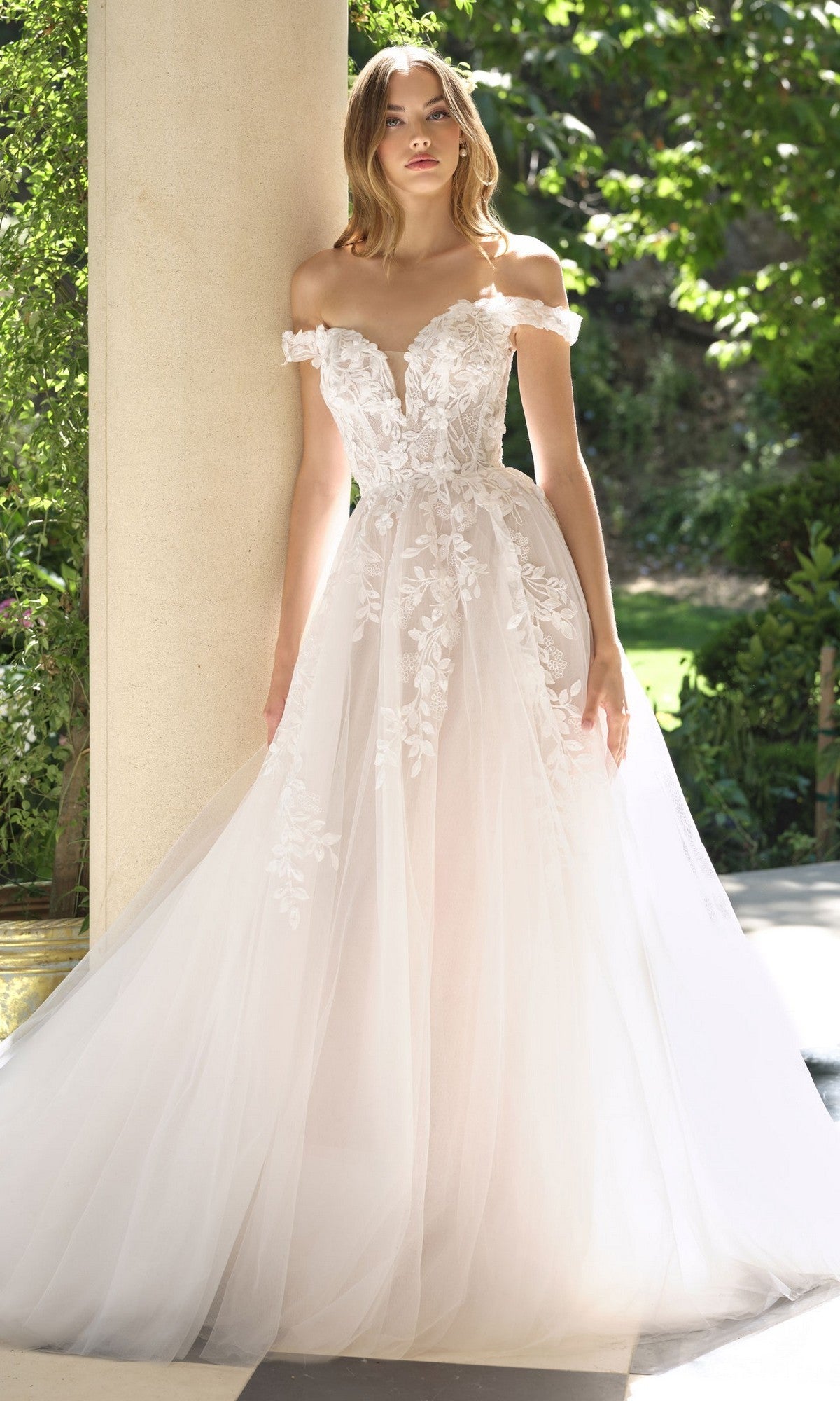 A woman stands outdoors wearing the Andrea and Leo WL057 long wedding dress in off white/nude, featuring off-shoulder floral lace details and a full A-line tulle skirt.